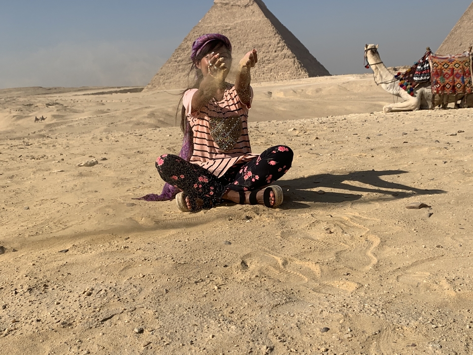 Personne assise sur le sable avec un chameau en arrière-plan et des pyramides visibles.