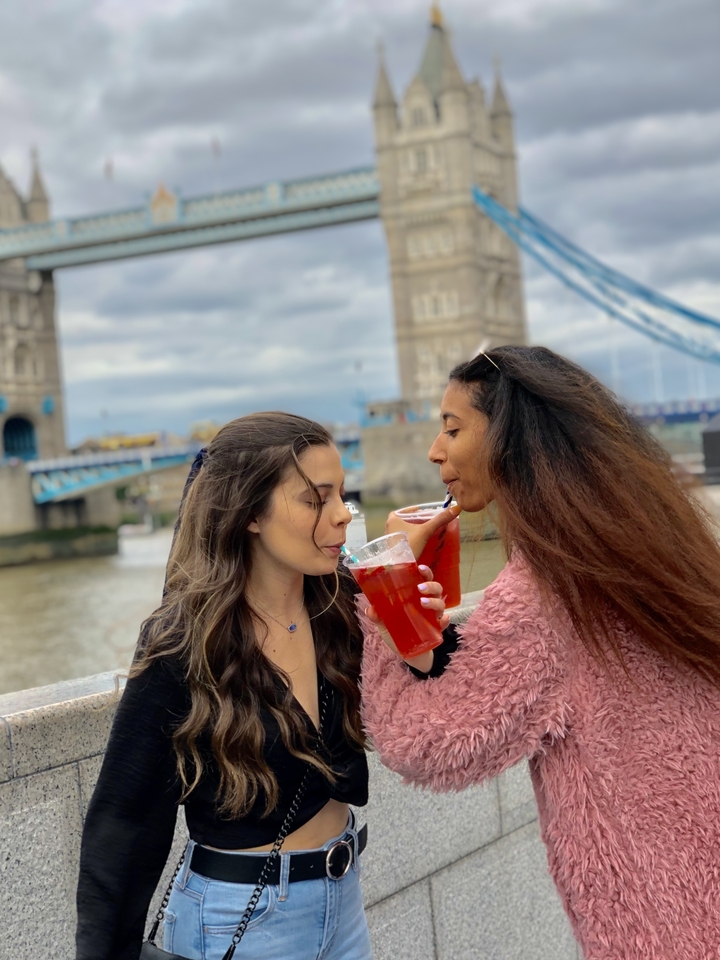 Two women enjoying drinks with a bridge and water in the background.