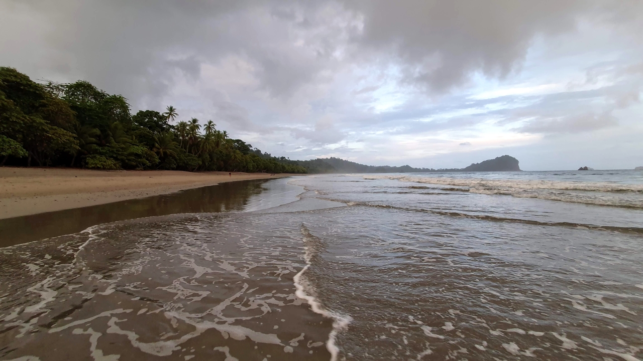 Scenic beach with waves and distant hills under a cloudy sky.