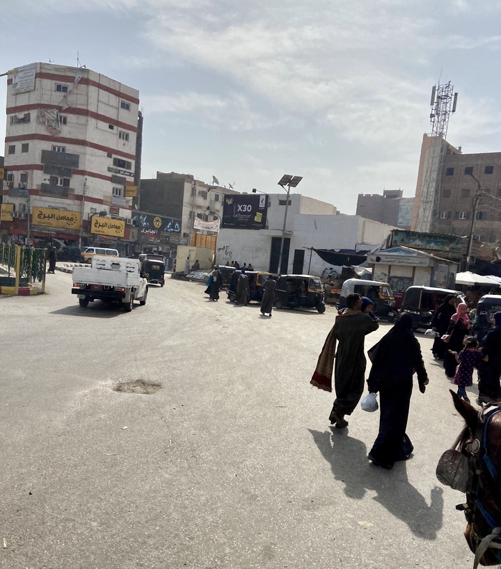 Bustling Egyptian street scene with tuk-tuks, pedestrians, and weathered shopfronts under hazy daylight.