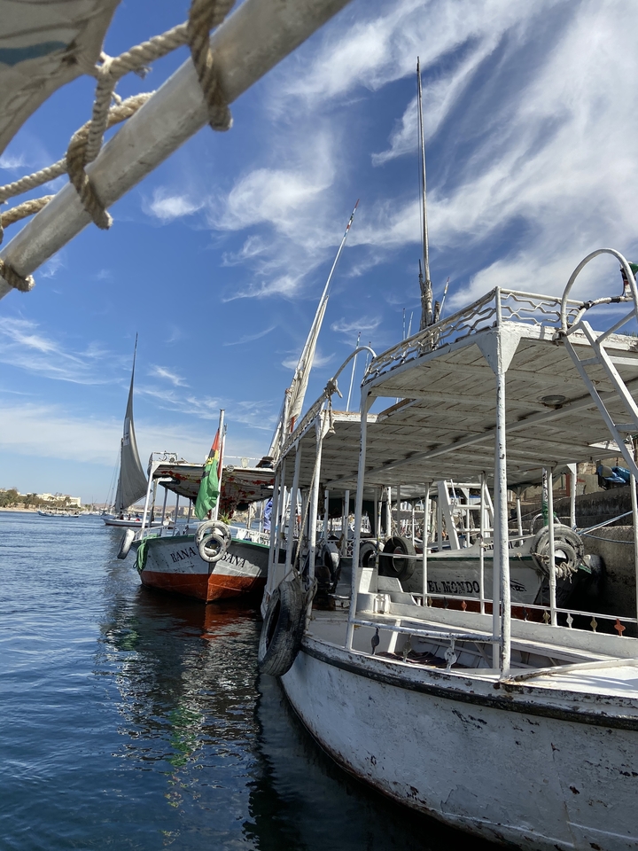 Row of moored feluccas under a vivid blue sky with wispy clouds along the banks of the Nile.