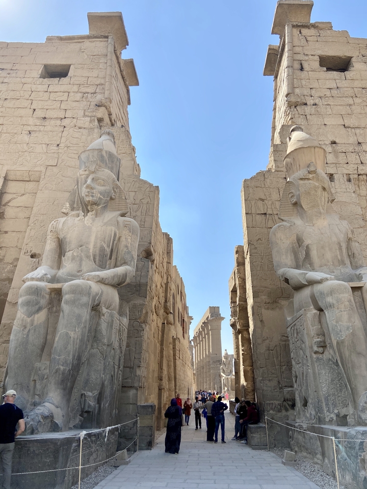 Twin colossal seated statues guard the entrance corridor of Luxor Temple against a clear blue sky.