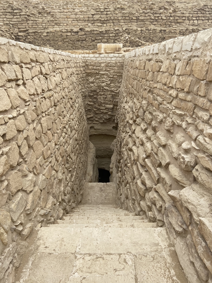 Narrow stone passageway with rough masonry walls descends into an ancient subterranean chamber.