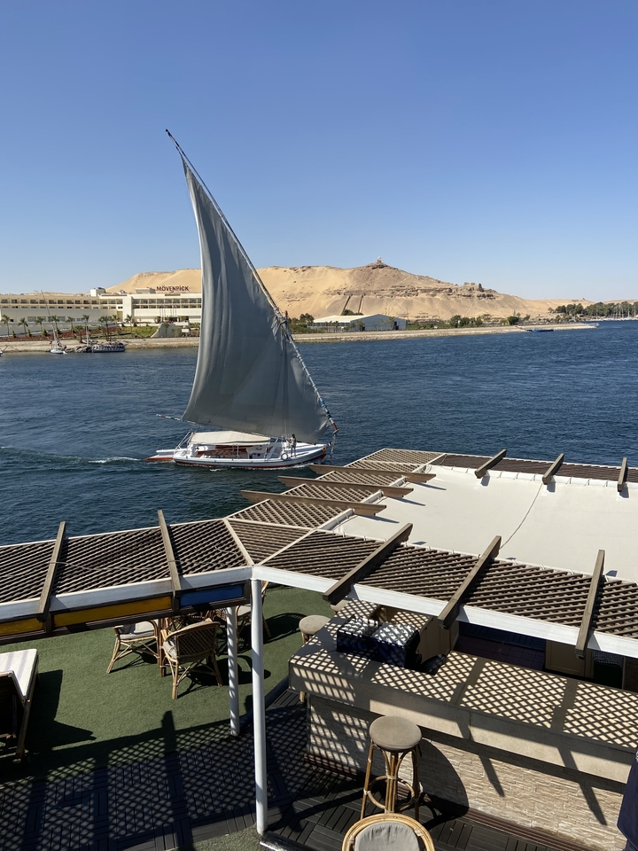 Traditional felucca with a tall triangular sail glides along the Nile with sandy hills and hotels behind.