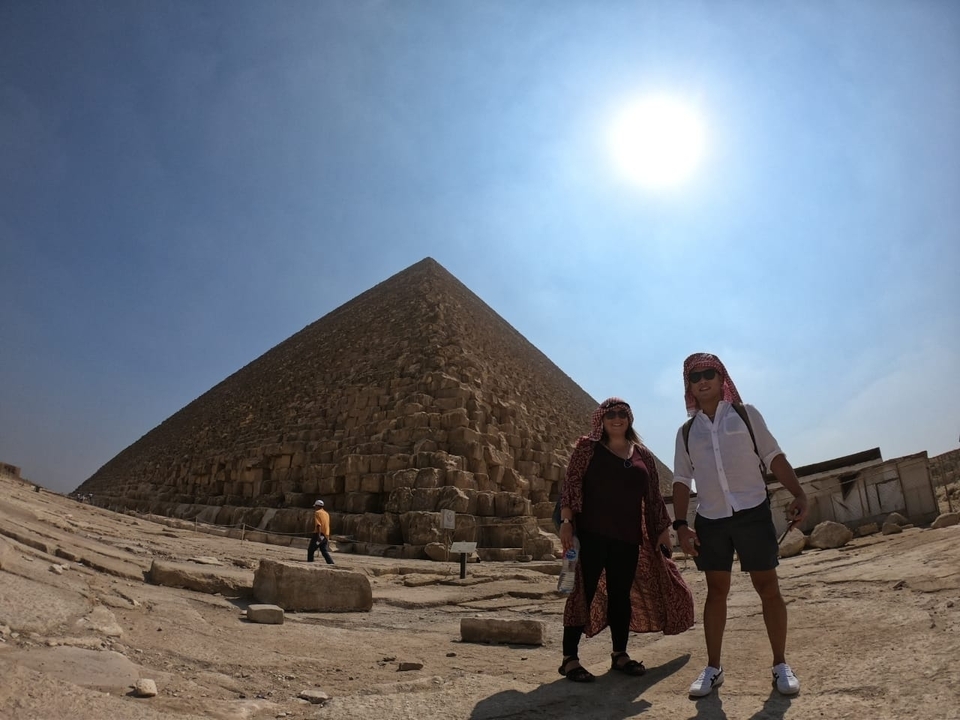 Two people standing in front of a large pyramid with bright skies.