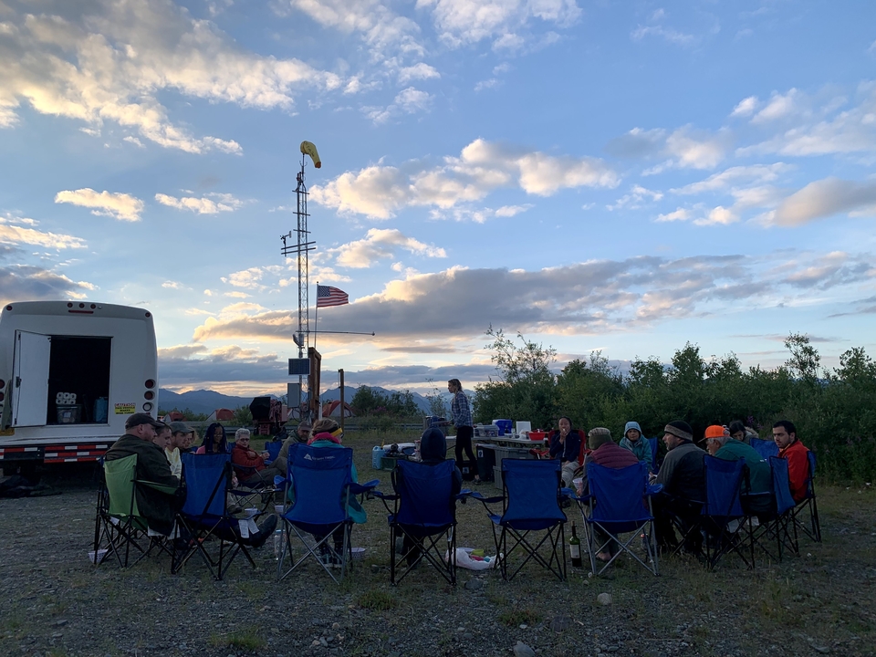 Group of people sitting in camping chairs near a bus with mountains in the background.