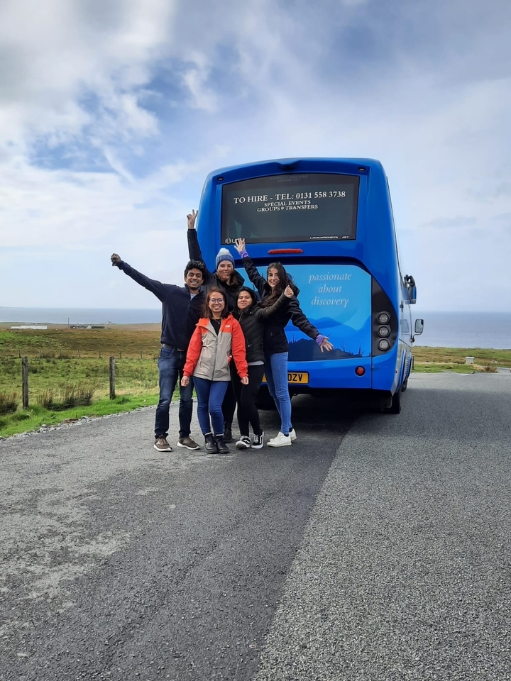 Group of friends posing with excitement next to a tour bus in a scenic rural area.