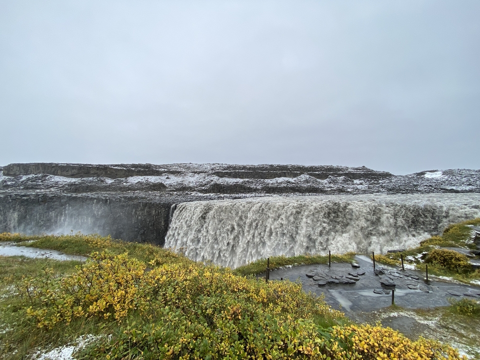 Massive waterfall flowing over a cliff, surrounded by snow and grass.