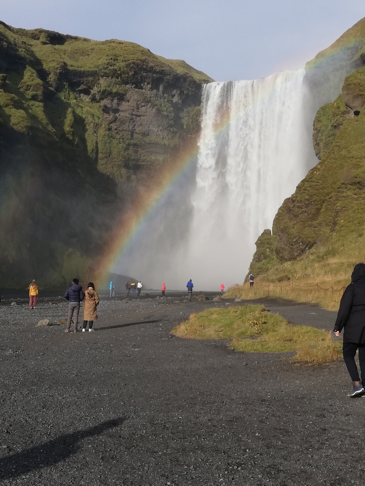 Rainbow over a waterfall with tourists standing nearby.