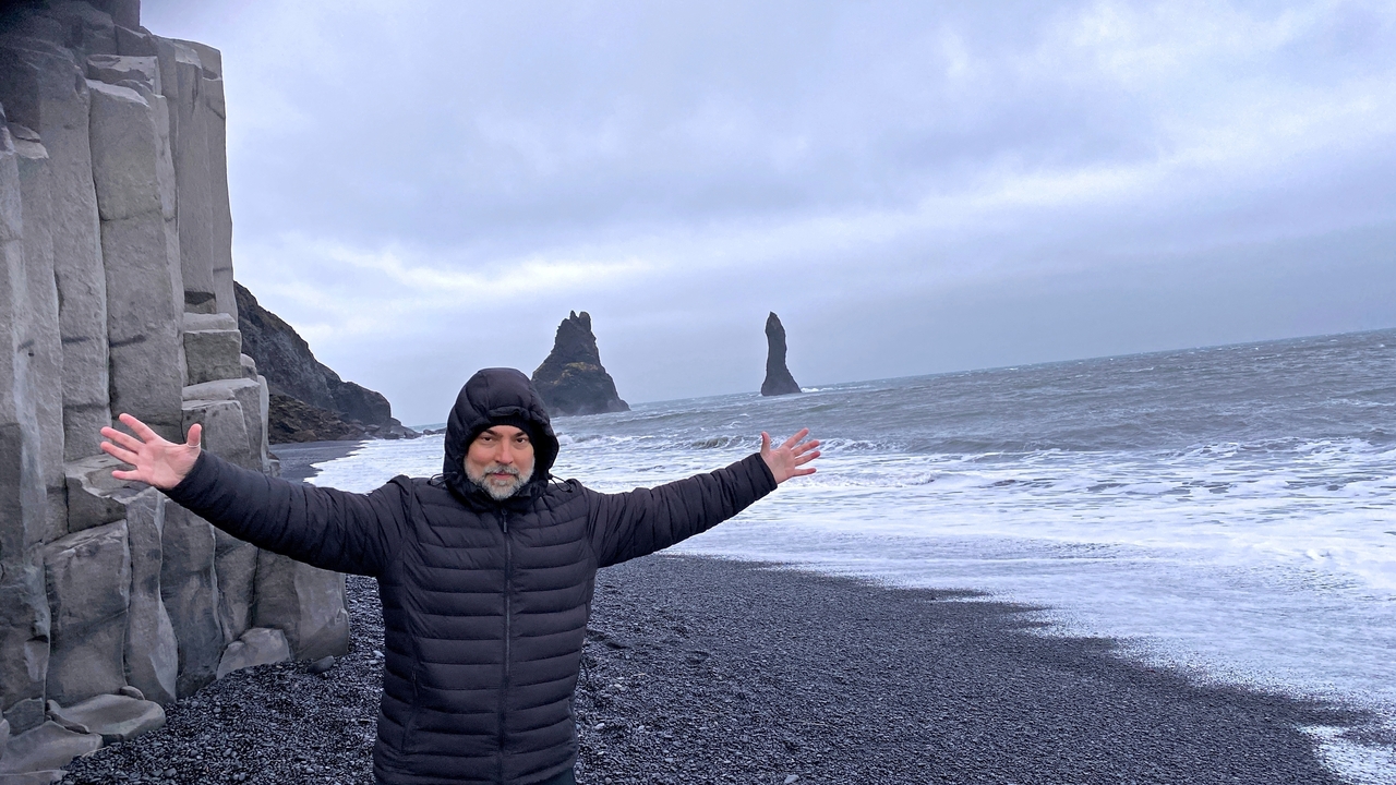 Person on a black sand beach with sea stacks in the background.