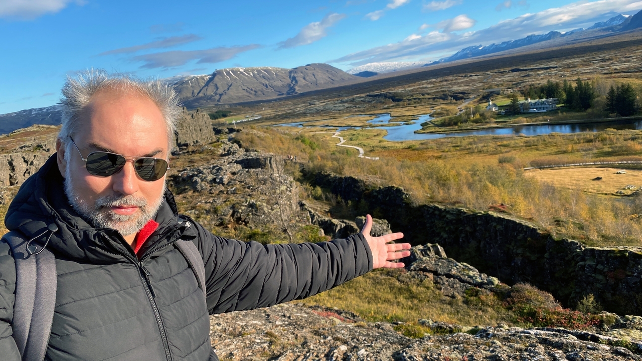 A man gesturing while standing on a rocky terrain with a scenic view.