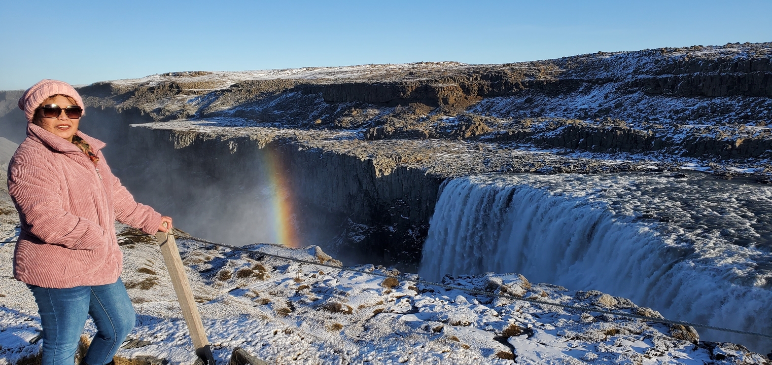 Person standing by a waterfall with a rainbow visible.