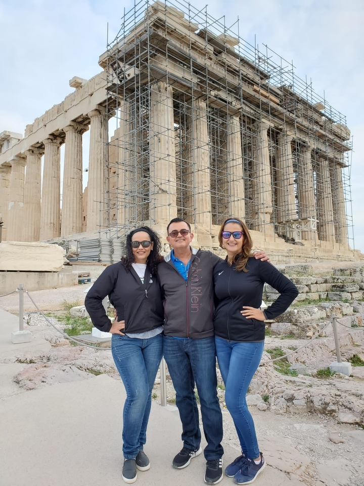 Trois personnes posent devant le Parthénon à Athènes avec des échafaudages visibles.