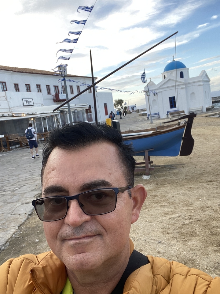 Un homme qui prend un selfie avec un bateau grec traditionnel en arrière-plan.
