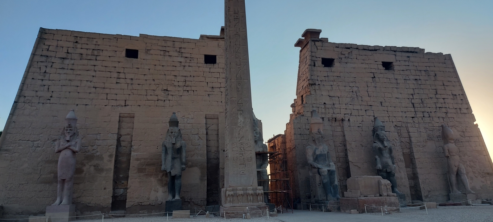 Ancient temple ruins with statues and an obelisk at dusk.