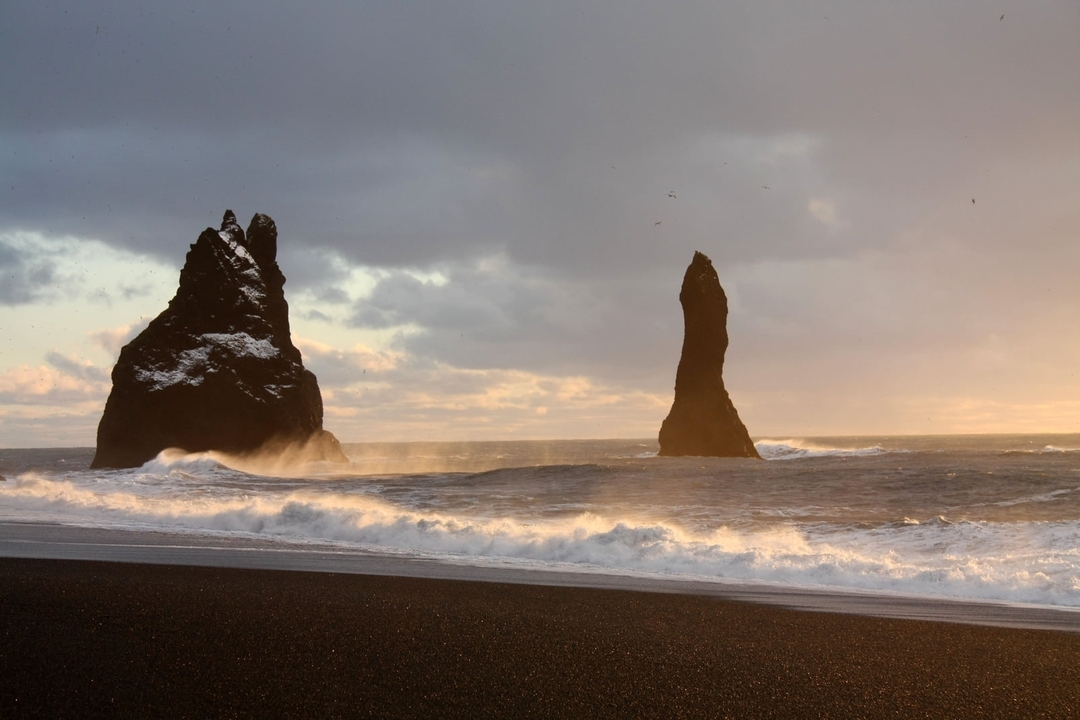 Piles rocheuses spectaculaires sur une plage de sable noir au coucher du soleil.