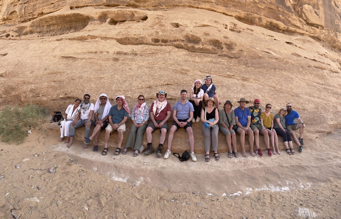 Group of people sitting against a rock wall in the desert.