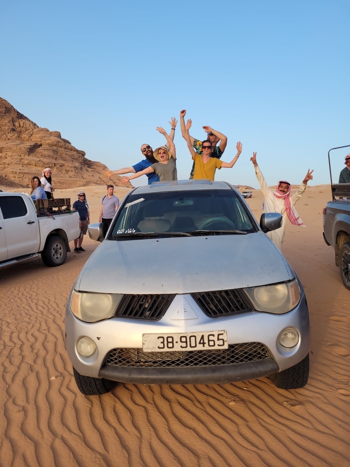 Group of people posing with arms raised next to a car in the desert.