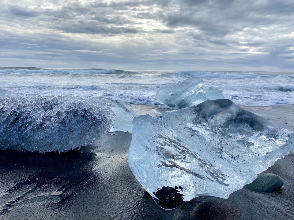 Clear ice on black sand beach with ocean waves.
