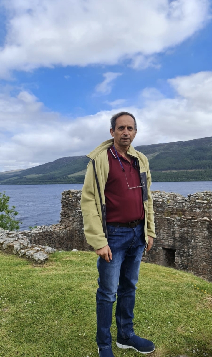 A person posing by a stone wall with a lake and hills in the background.