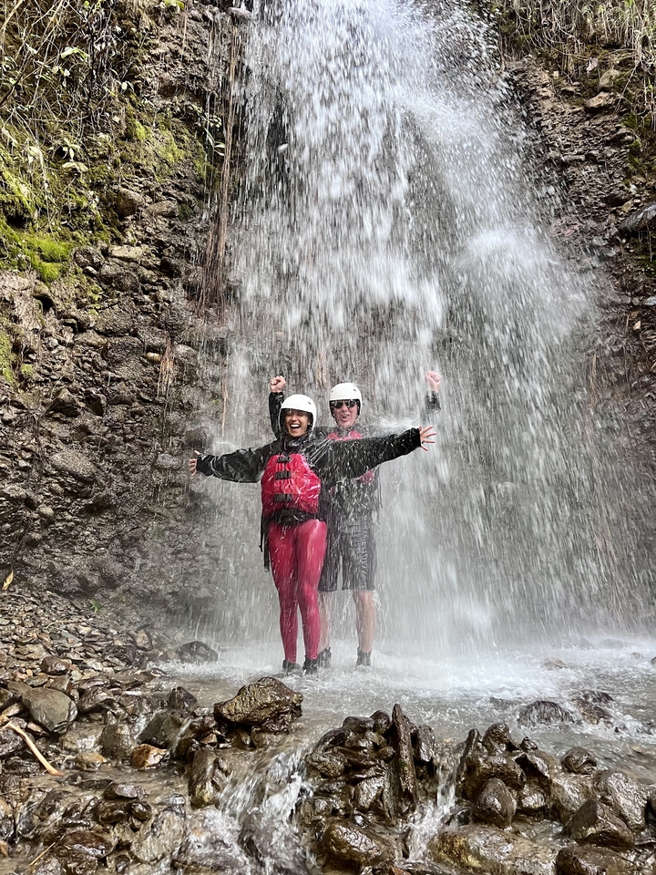 Couple under a waterfall, wearing helmets and life vests, with joyful expressions.