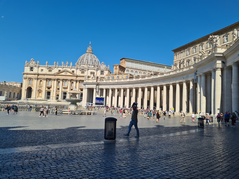 A busy square with people in front of a large basilica and colonnade.