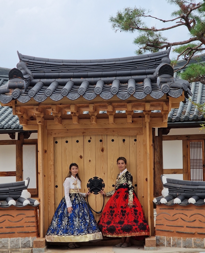 Two people in traditional attire standing in front of a wooden gate.