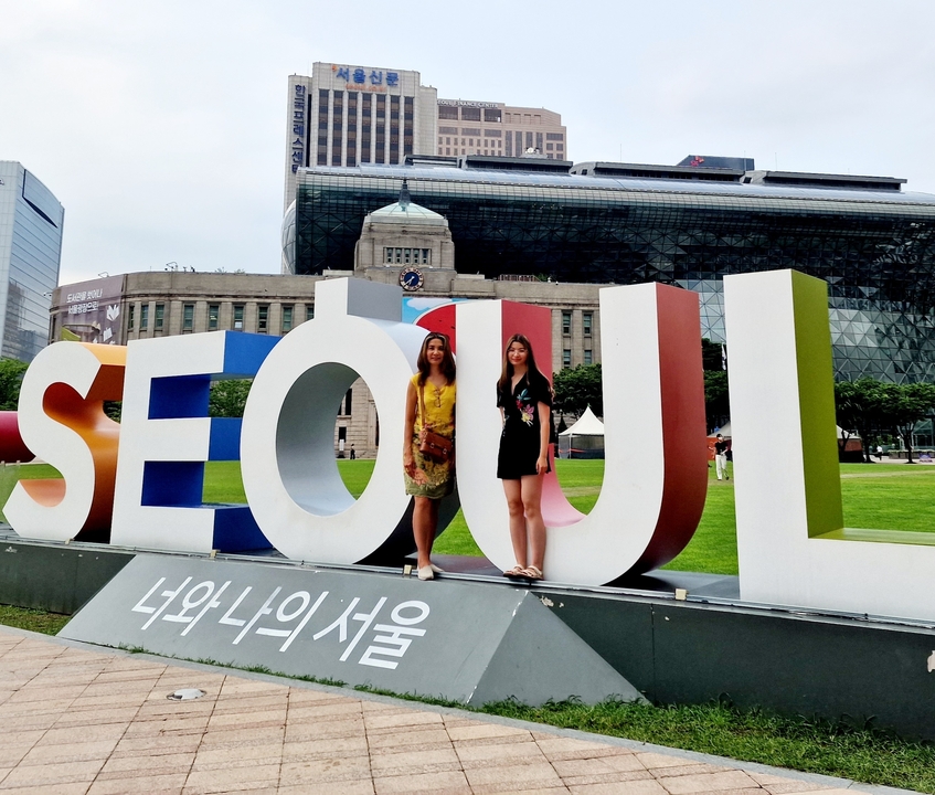 Two people standing in front of a large 'SEOUL' sign in a park.