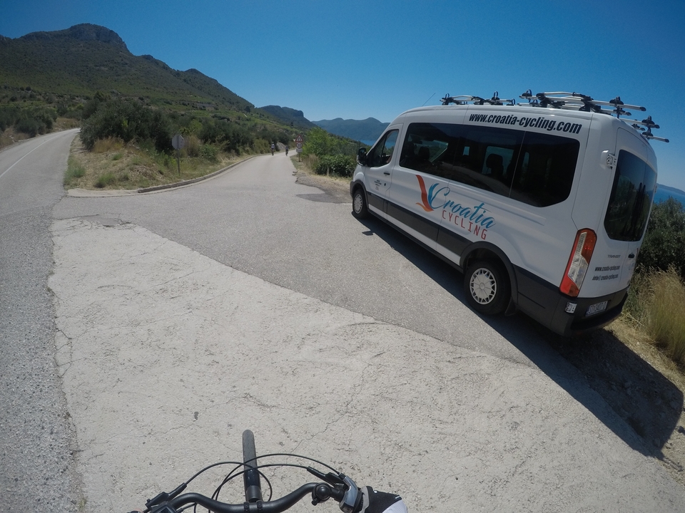 Cycling support van parked along a scenic road with a view of mountains and the sea.