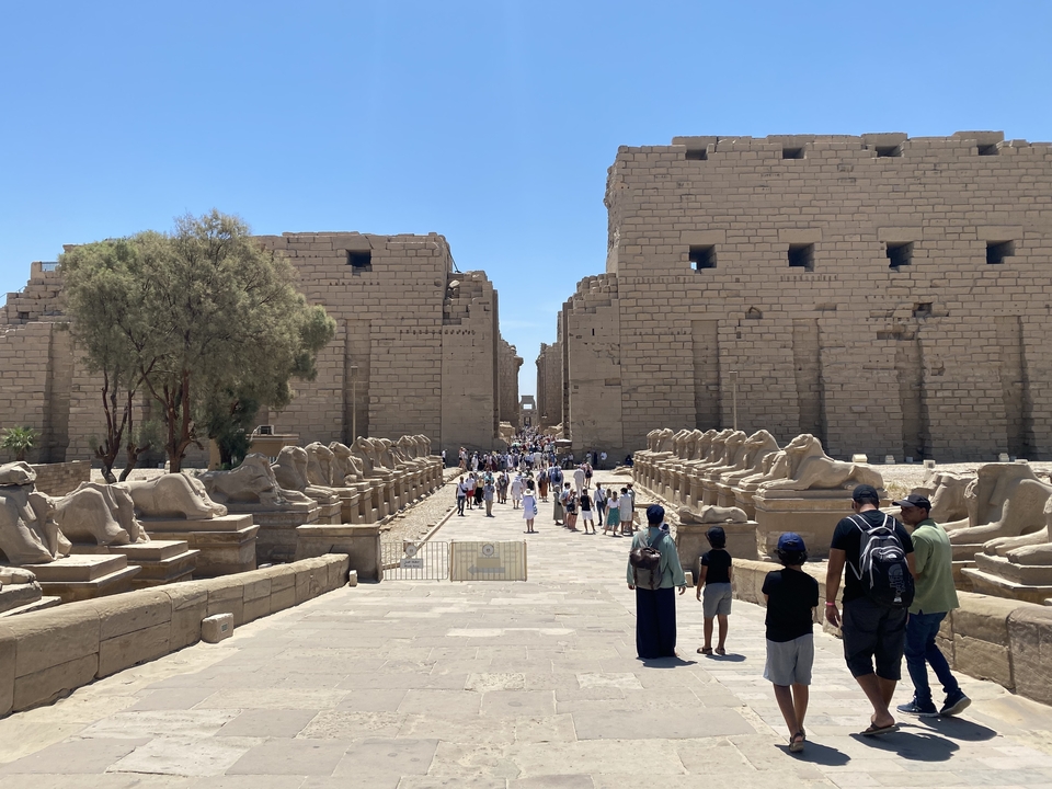 Avenue de sphinx menant à l'entrée d'un temple, avec des visiteurs qui marchent.