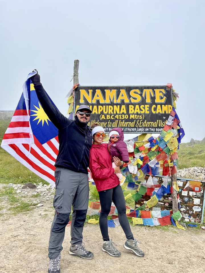 Une famille posant avec un drapeau devant le panneau du camp de base de l'Annapurna, entourée de drapeaux de prière.
