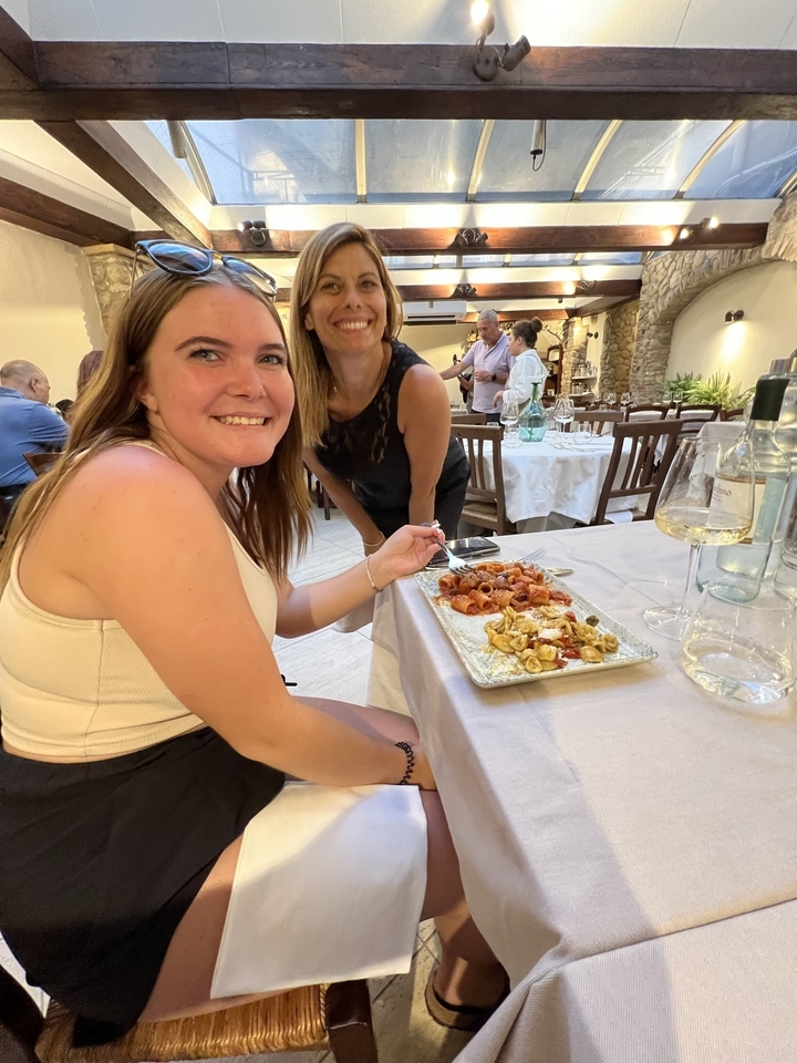 Two women smiling at a table with an array of pasta dishes.