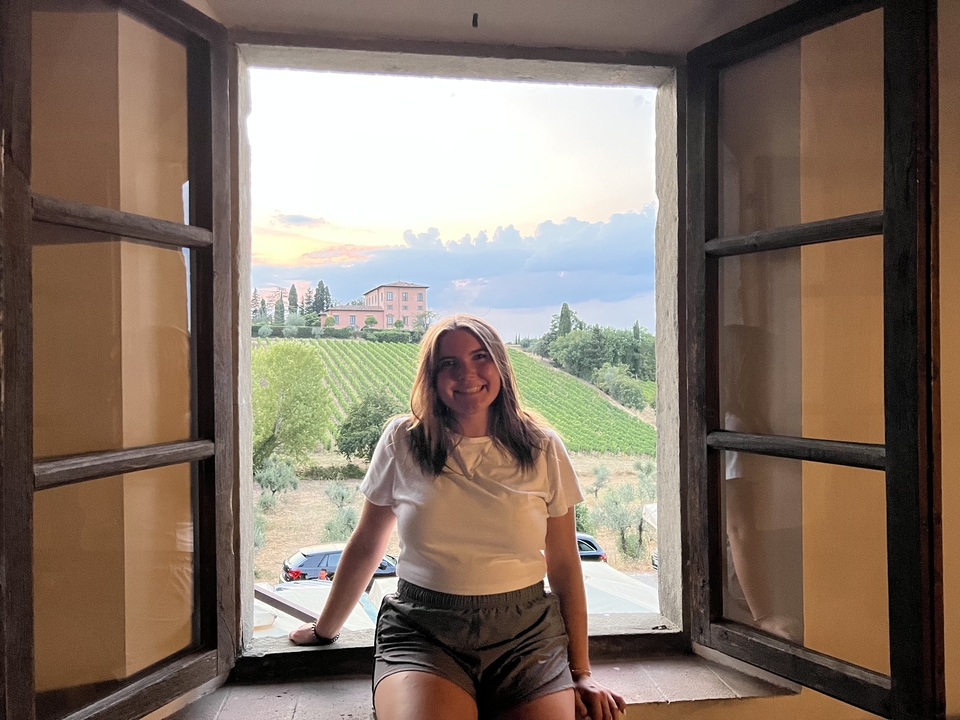 Woman posing by a window with a sunset view over vineyards.