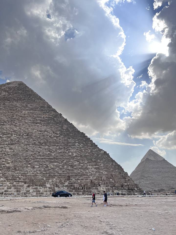 Pyramid under a dramatic sky with some visitors and a car.