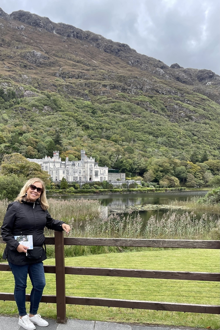 Woman posing in front of a scenic castle with greenery.