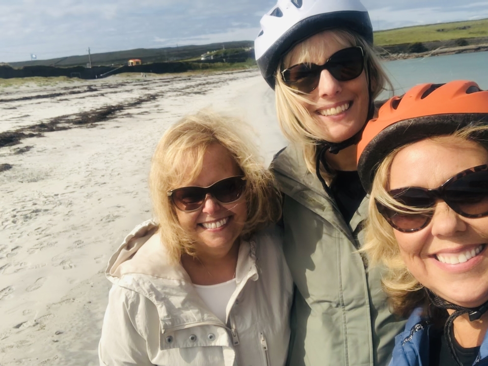 Trois femmes souriantes avec des vélos sur une plage.