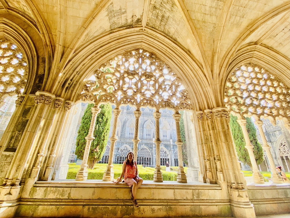 Person sitting under an ornate stone archway with a garden and building in the background.
