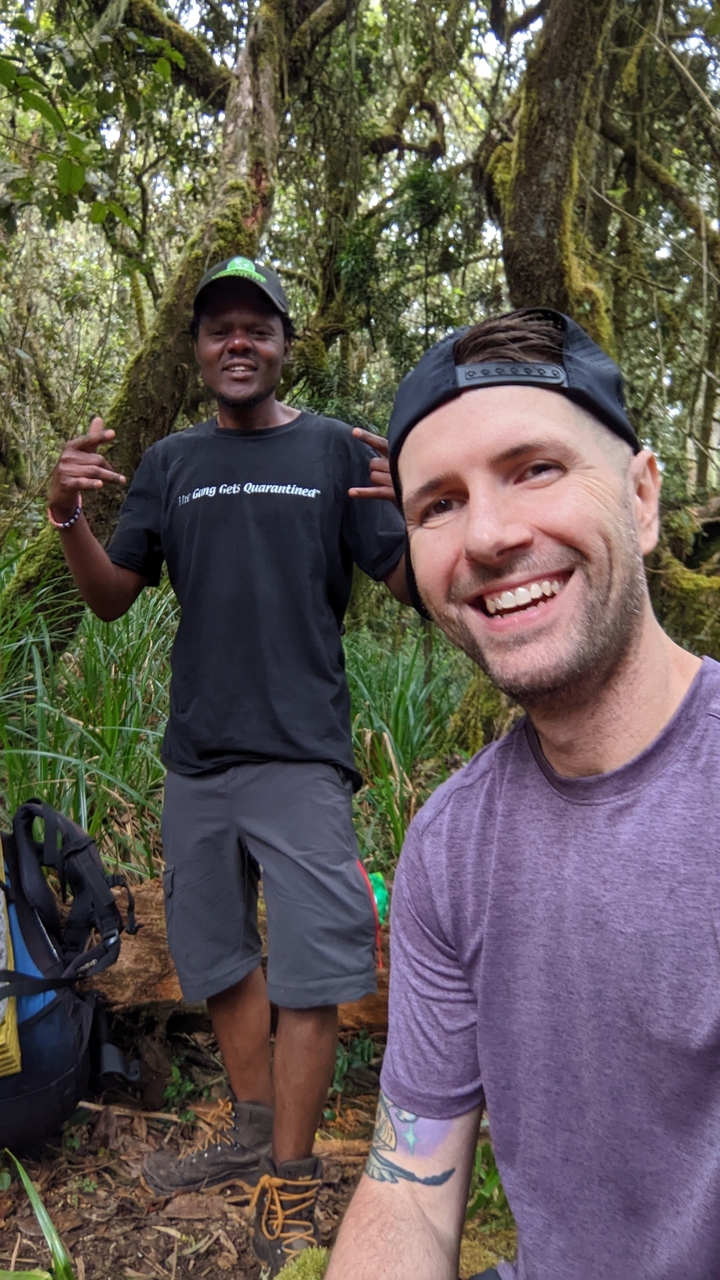 Two men smiling in a forested area showing peace sign.
