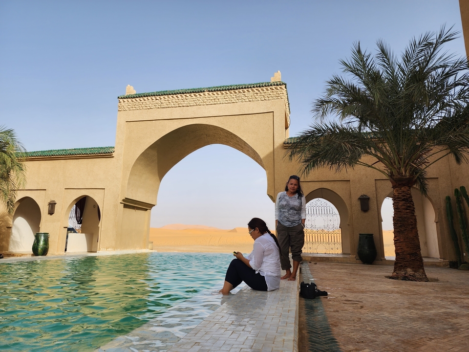 Two people by a pool under an archway in a desert locale.
