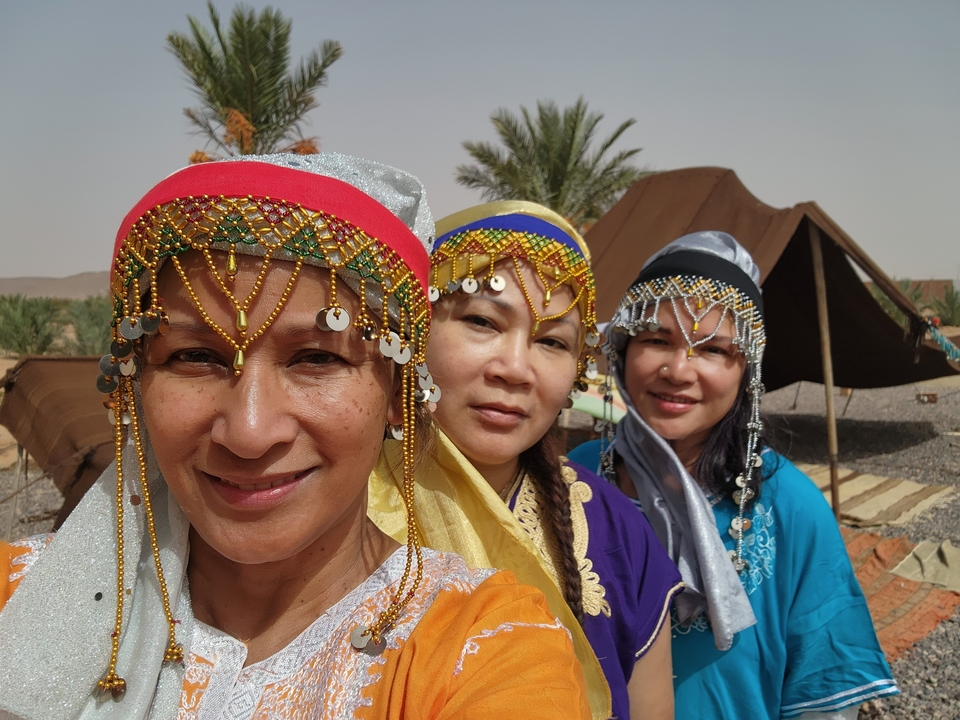 Three women in traditional attire with tents in the background.