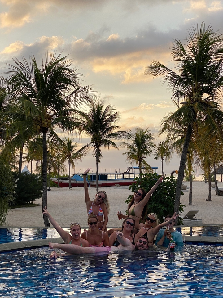 Group of people joyfully posing on a beach with palm trees and a boat in the background.