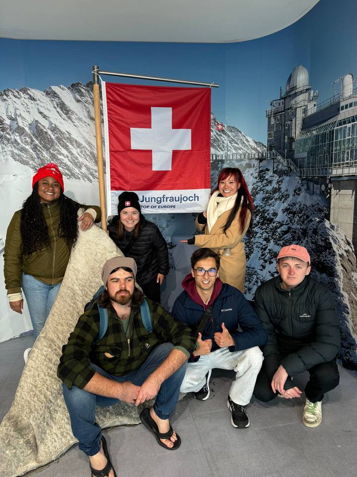 Group posing under Swiss flag indoors.
