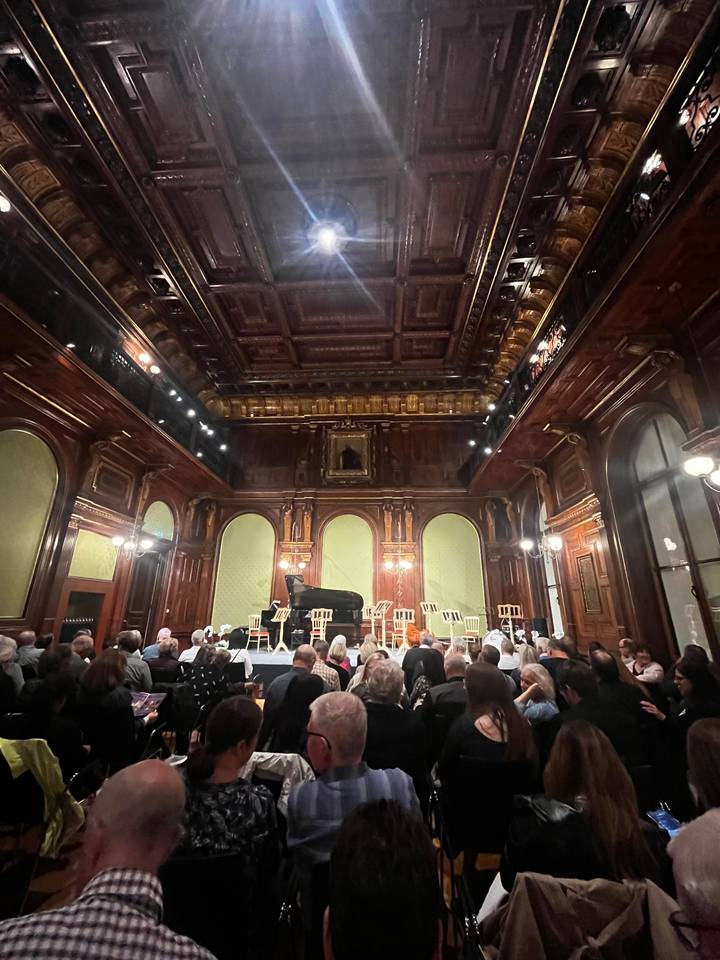 Audience inside a dark wooden-paneled concert hall.