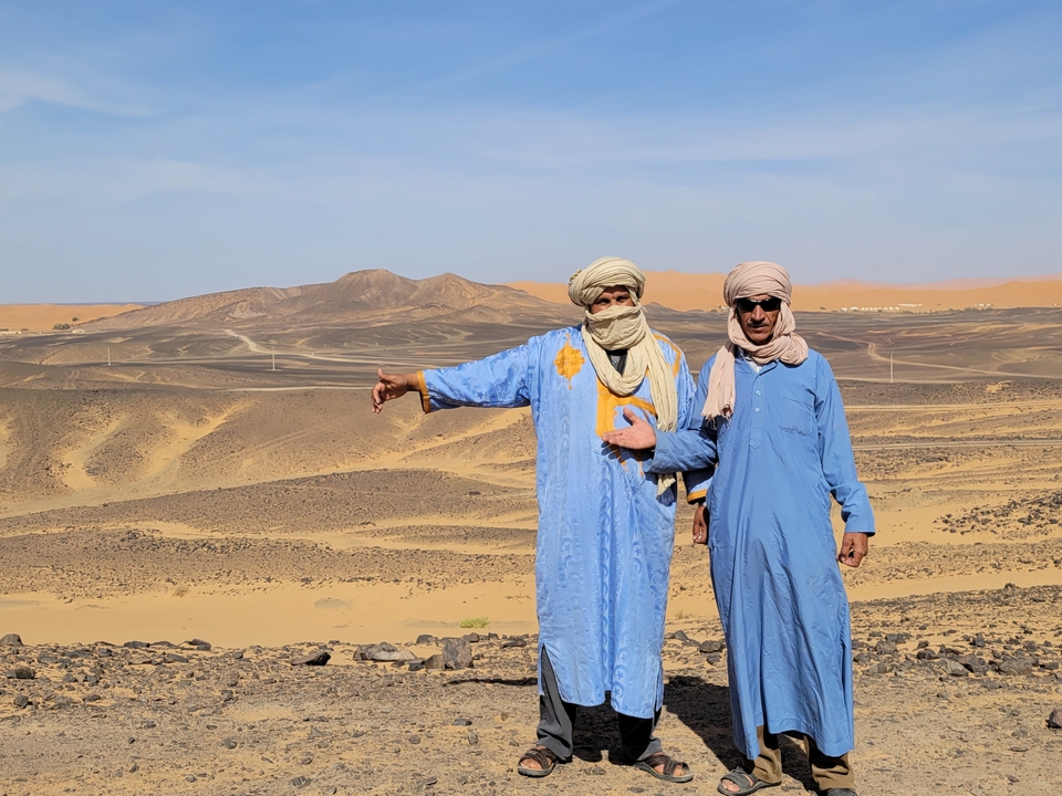 Two men in traditional attire gesturing towards a desert landscape.