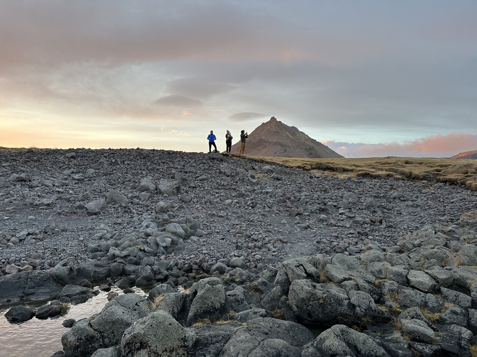 Three people standing on a rocky terrain with a mountain in the background.