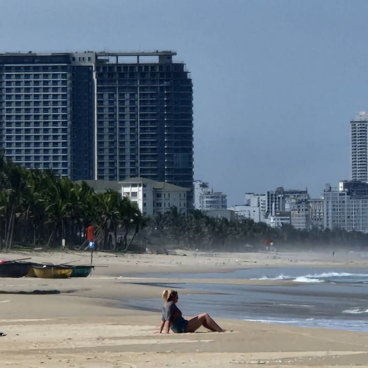 People sitting on beach with city and trees in background.