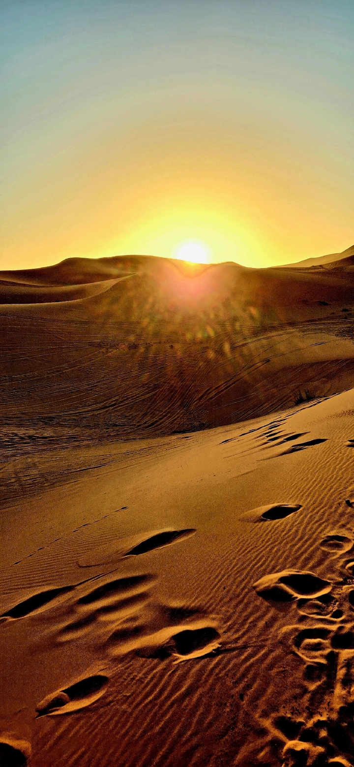 Sunlit sand dunes with visible tracks and a lens flare effect.