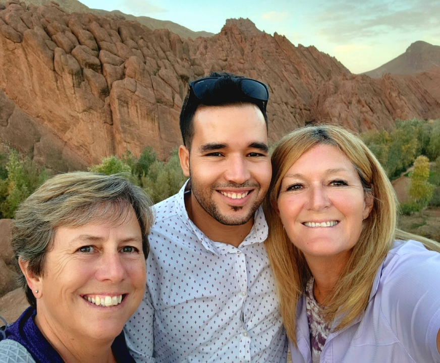 Three smiling people with sunglasses in front of a desert road and mountains.