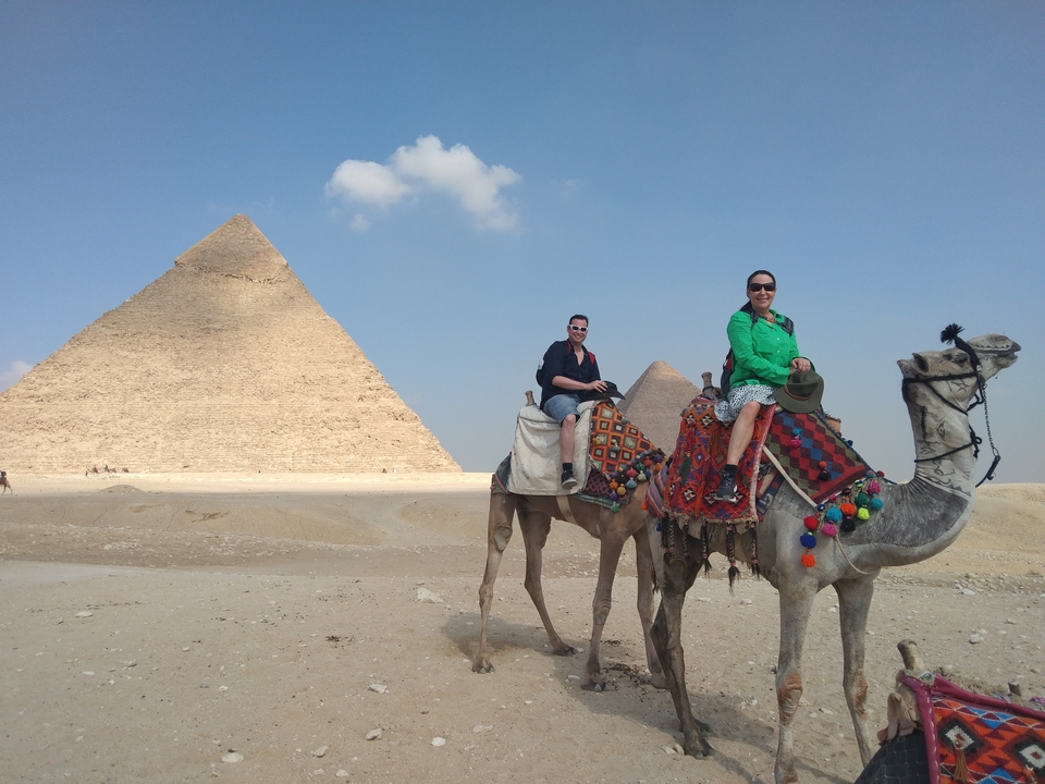 Two people in front of a pyramid on camels.