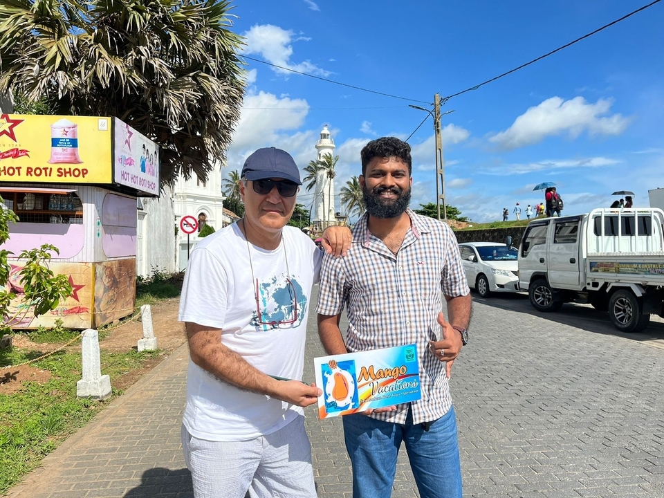 Two people standing on a street with a brightly colored sign reading 'Mango Madness'.